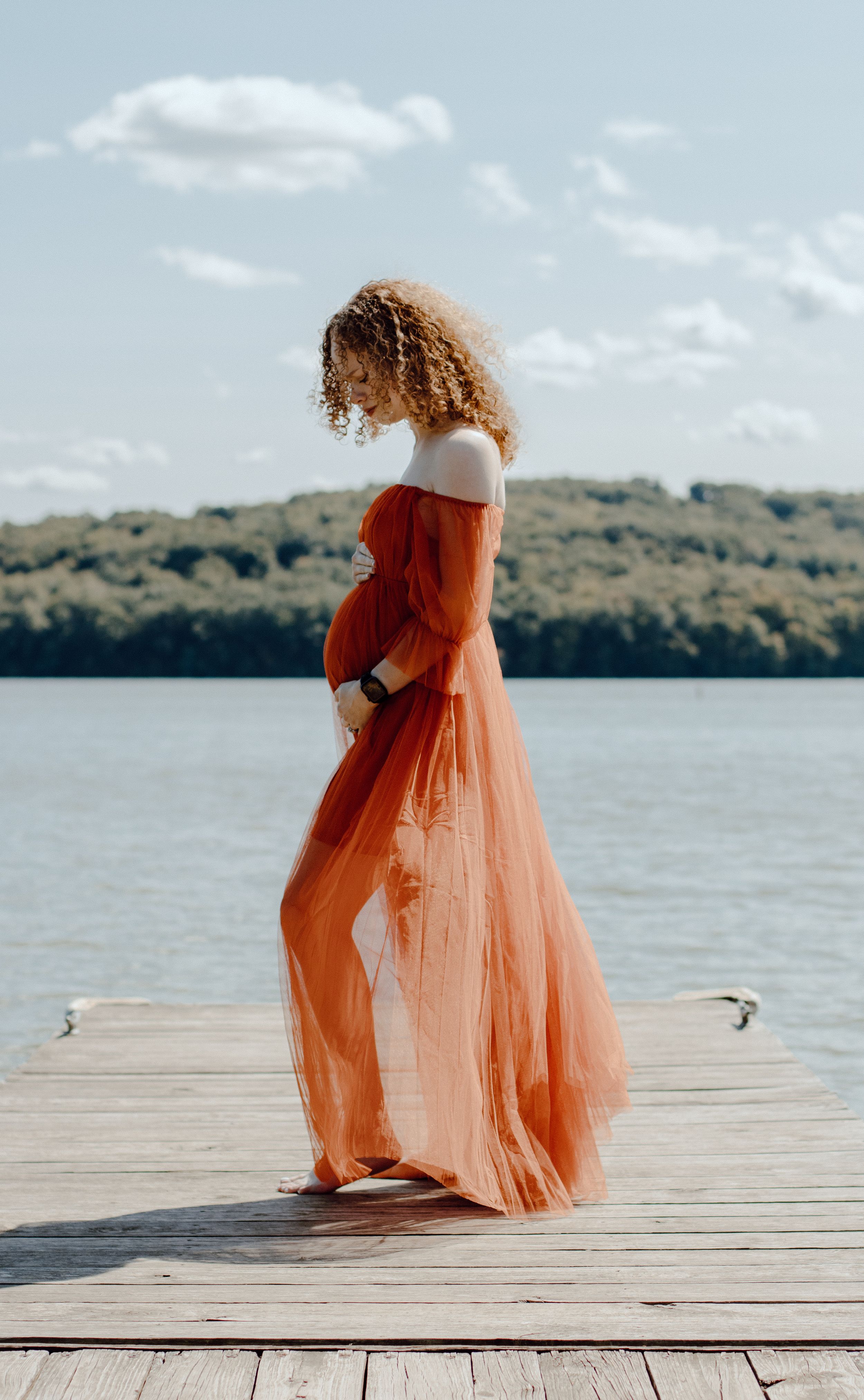 Side view of woman in flowing orange dress standing on wooden dock by lake.