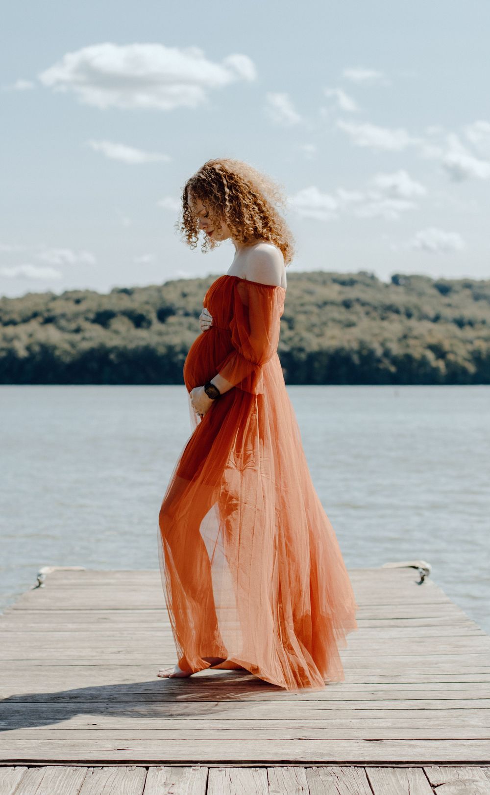 Side view of woman in flowing orange dress standing on wooden dock by lake.