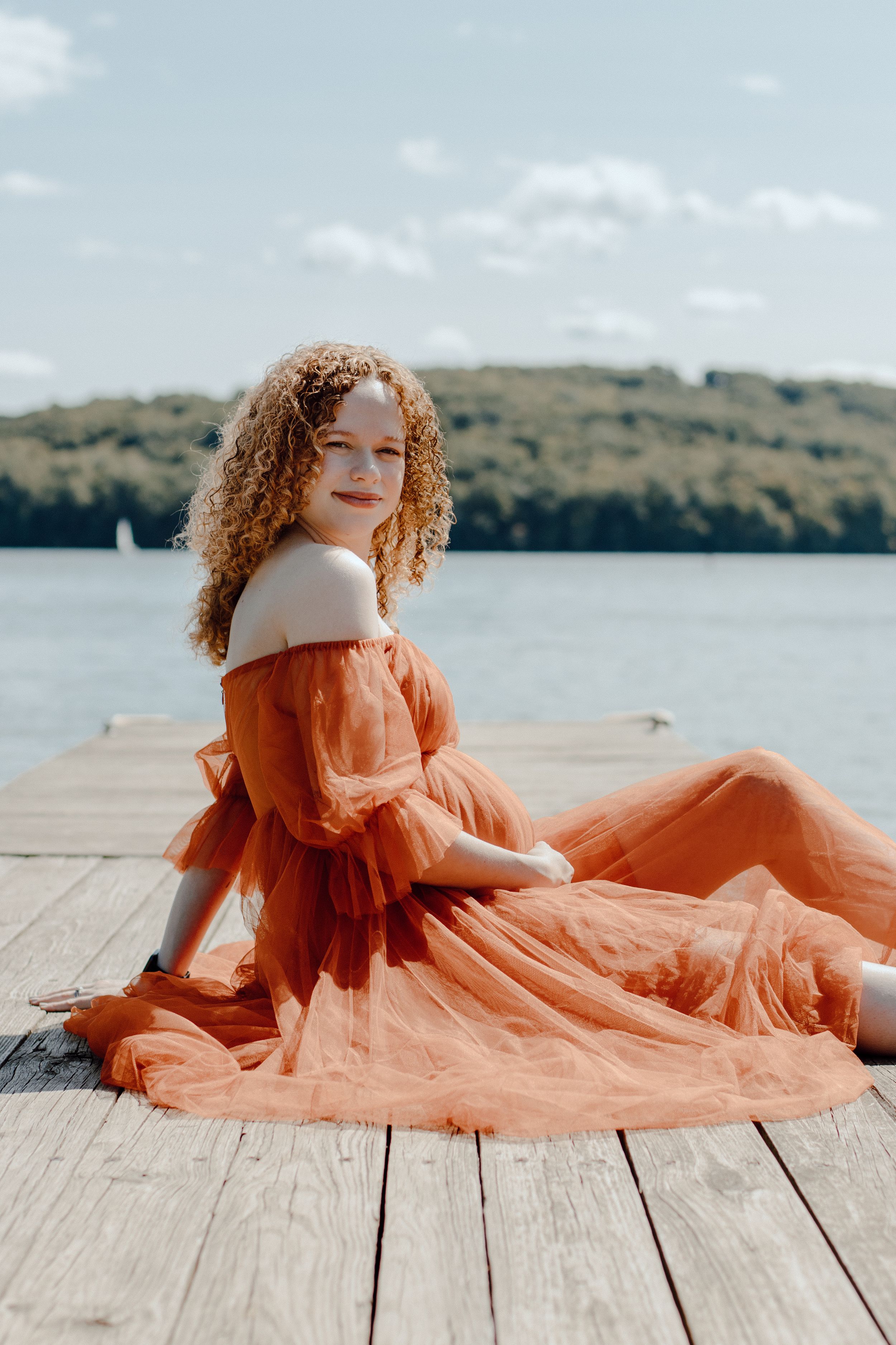 Woman in orange off-shoulder dress sitting on wooden dock by serene lake.