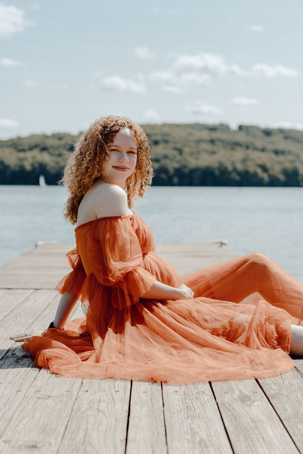 Woman in orange off-shoulder dress sitting on wooden dock by serene lake.