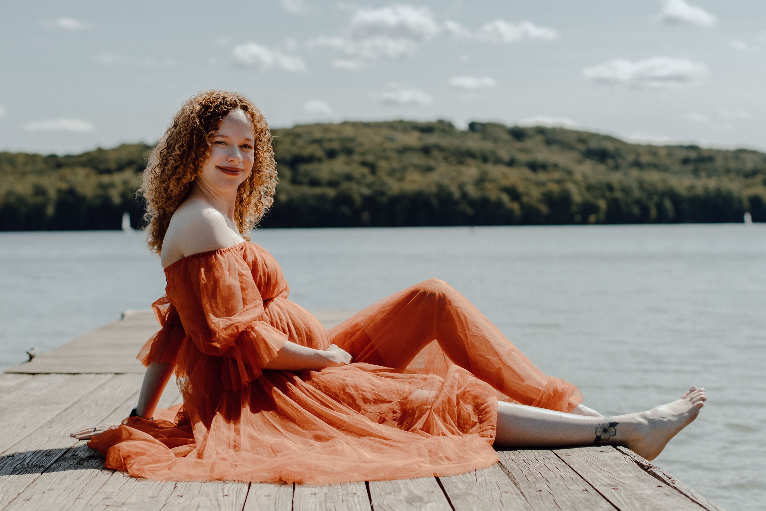 Woman in orange dress reclining on wooden dock with lake and hills in background.