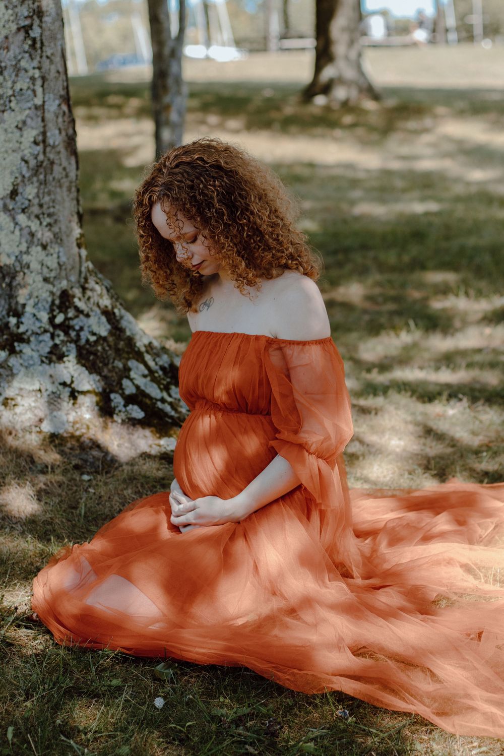 A flowing burnt orange dress creates an ethereal outdoor portrait against dappled sunlight and trees.