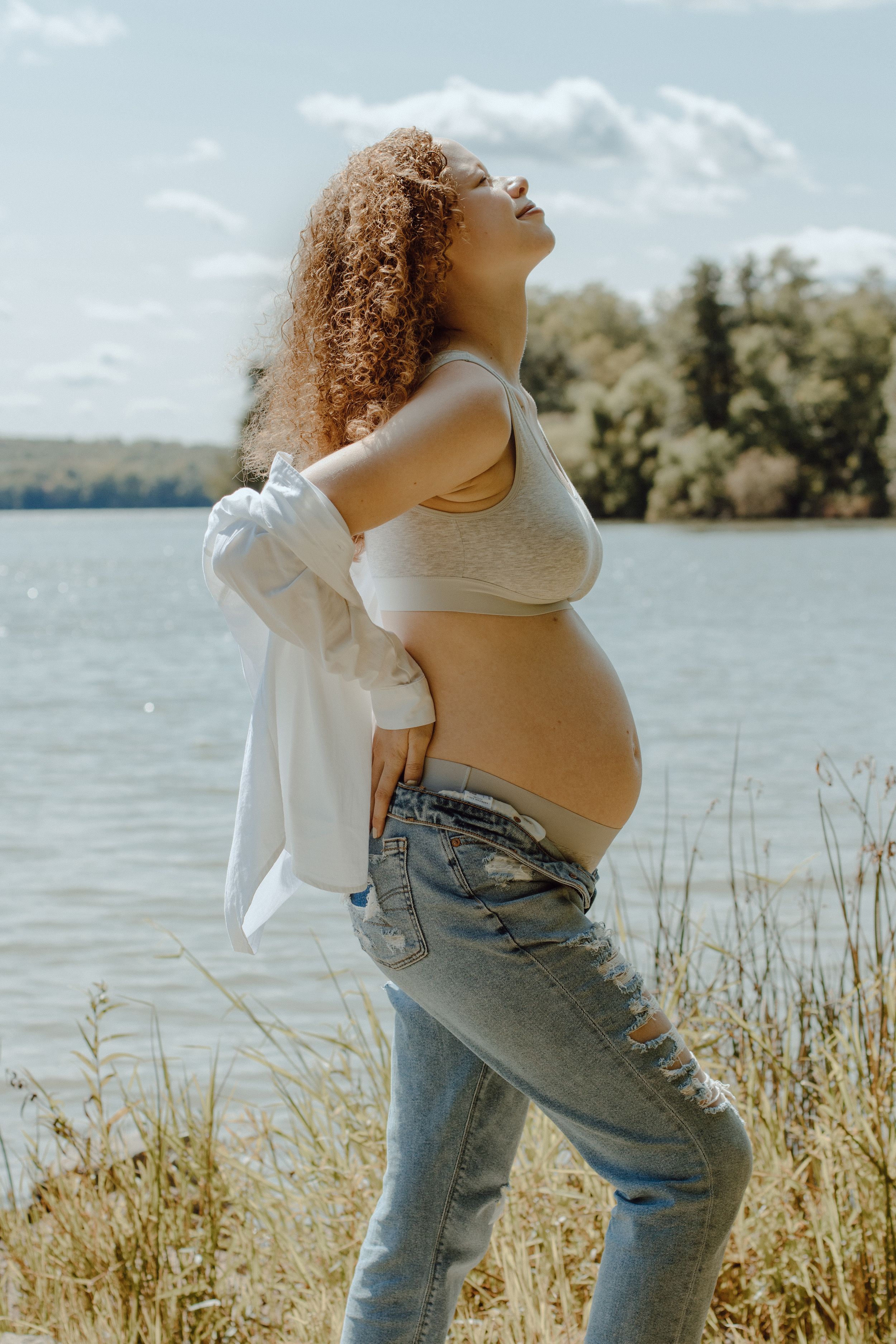 Person in casual attire showing pregnant belly against lakeside landscape.
