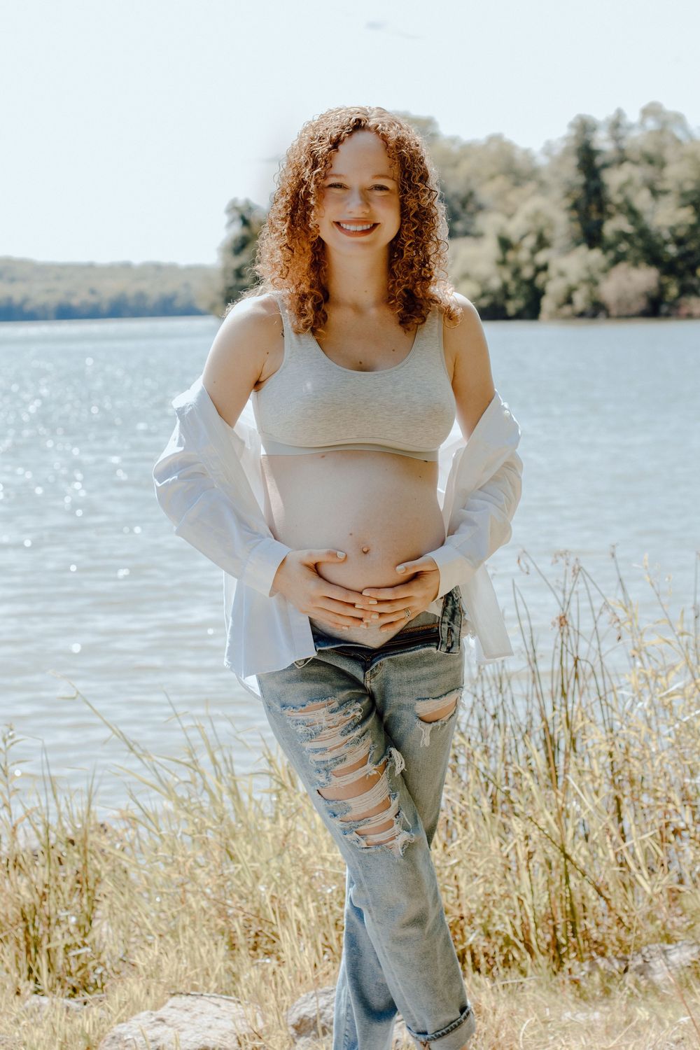 Sunny lakeside maternity photo with tall grass and water in background.