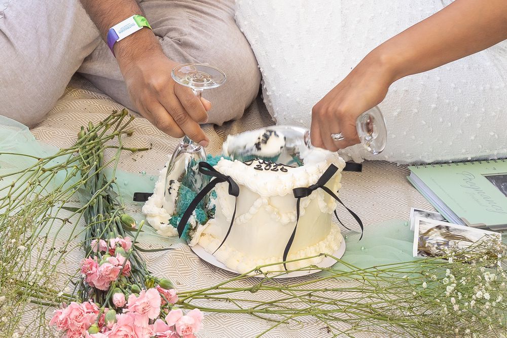 Close-up of hands breaking apart a white cake decorated with turquoise accents and surrounded by pink flowers.