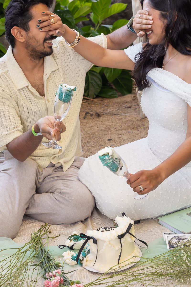 A couple playfully smashes cake on each other's faces during a celebration while holding turquoise drinks.