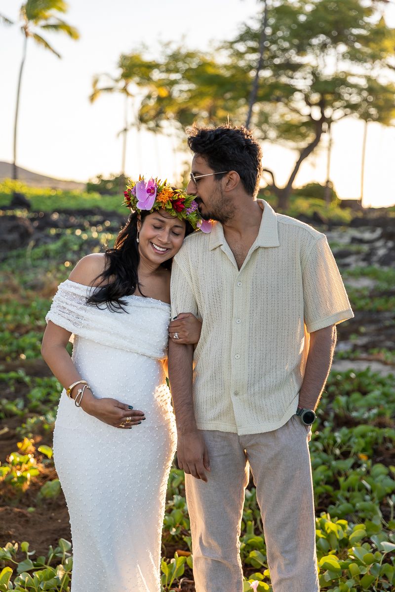 A romantic couple in light-colored clothing embraces outdoors with palm trees and greenery in the background at sunset.