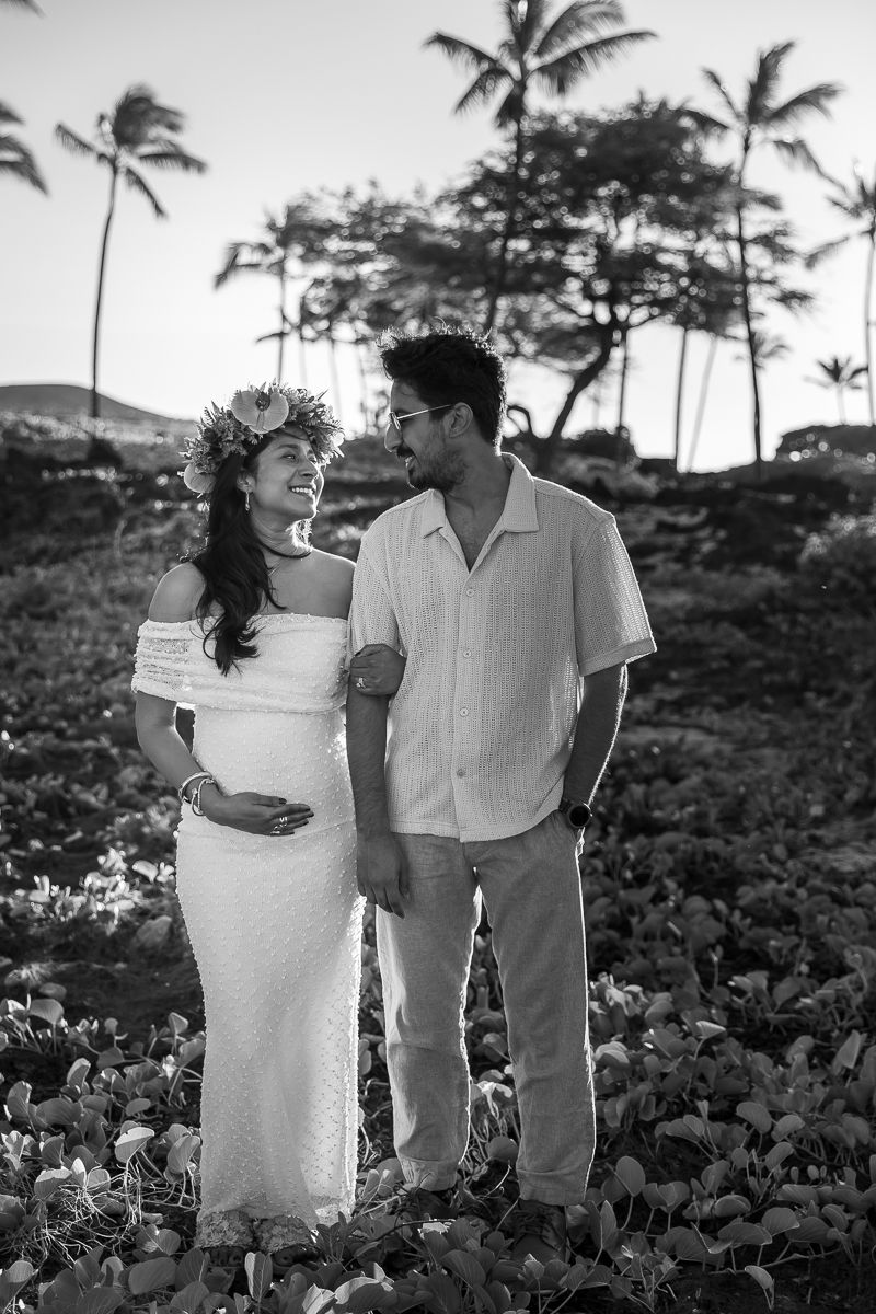 A black and white portrait of a couple in white attire standing together among tropical foliage and palm trees.