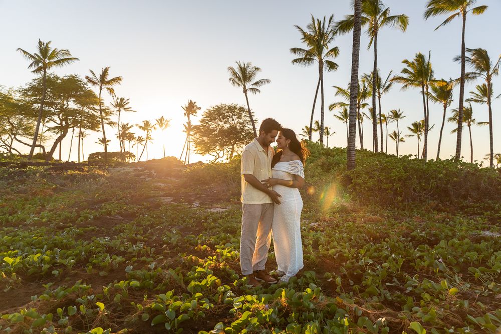A couple stands in a lush tropical garden with palm trees and golden sunlight at sunset in Hawaii.