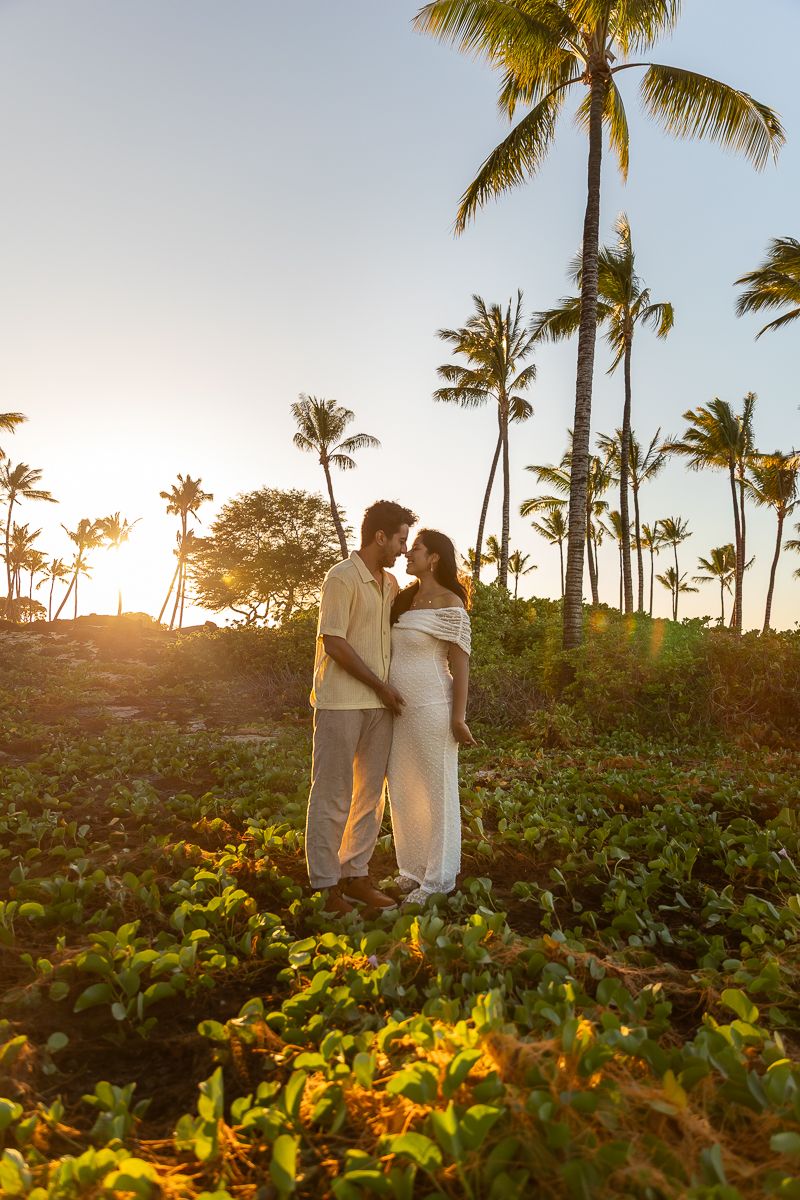 A romantic couple embraces among tall palm trees at sunset on a tropical hillside.