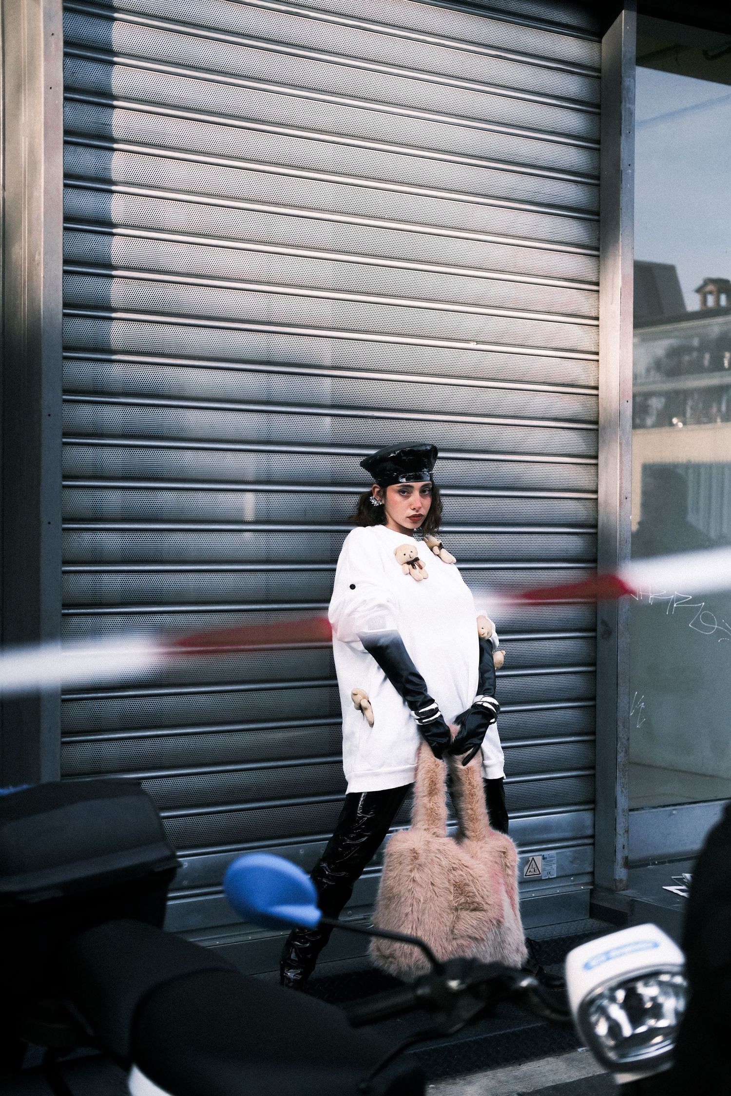 A person in a white top and fur skirt stands against an industrial metal shutter door.