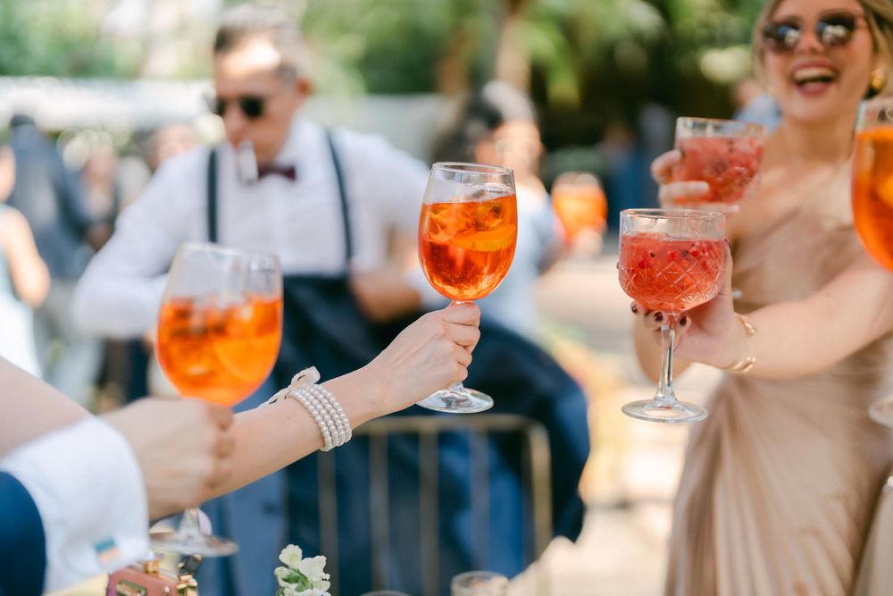 People holding orange Aperol Spritz cocktails in stemmed glasses at an outdoor social gathering.