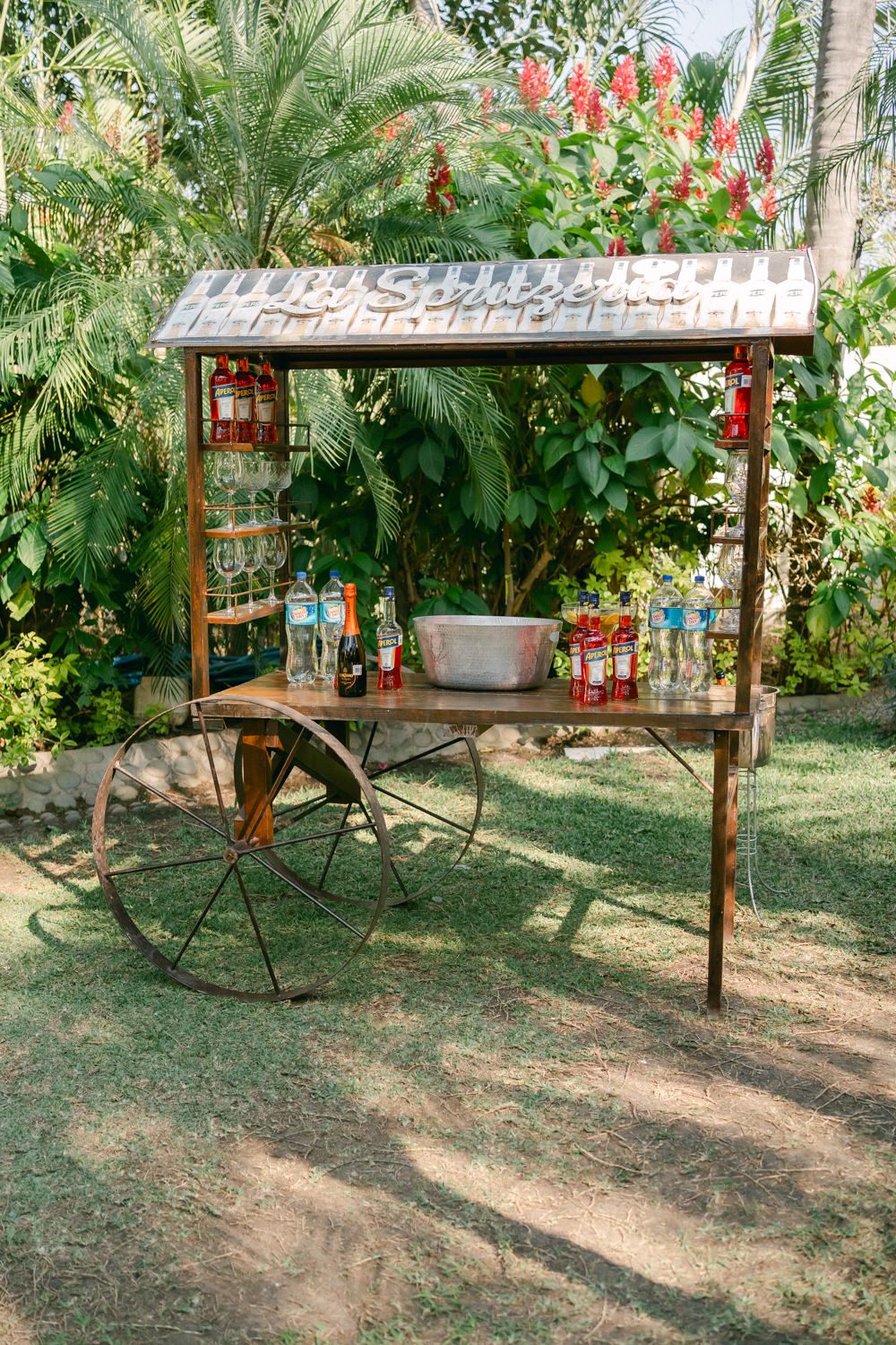 A rustic wooden pergola structure in a garden setting with red flowering plants and palm trees in the background.