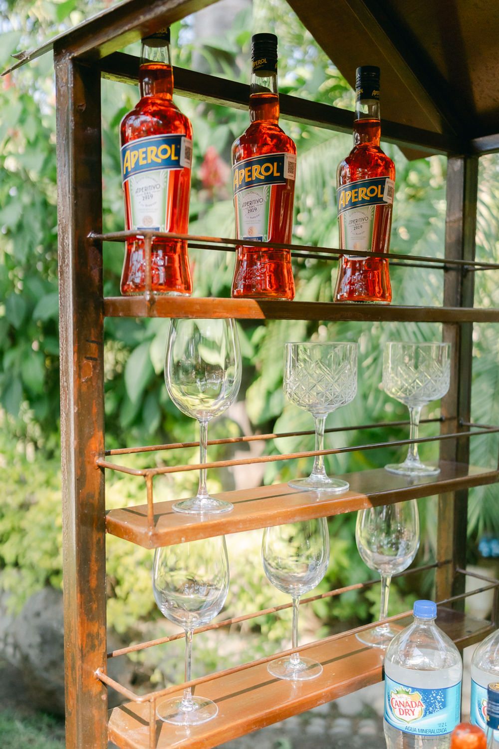 Wooden shelves displaying glass stemware and red liquor bottles arranged in an outdoor bar setting.