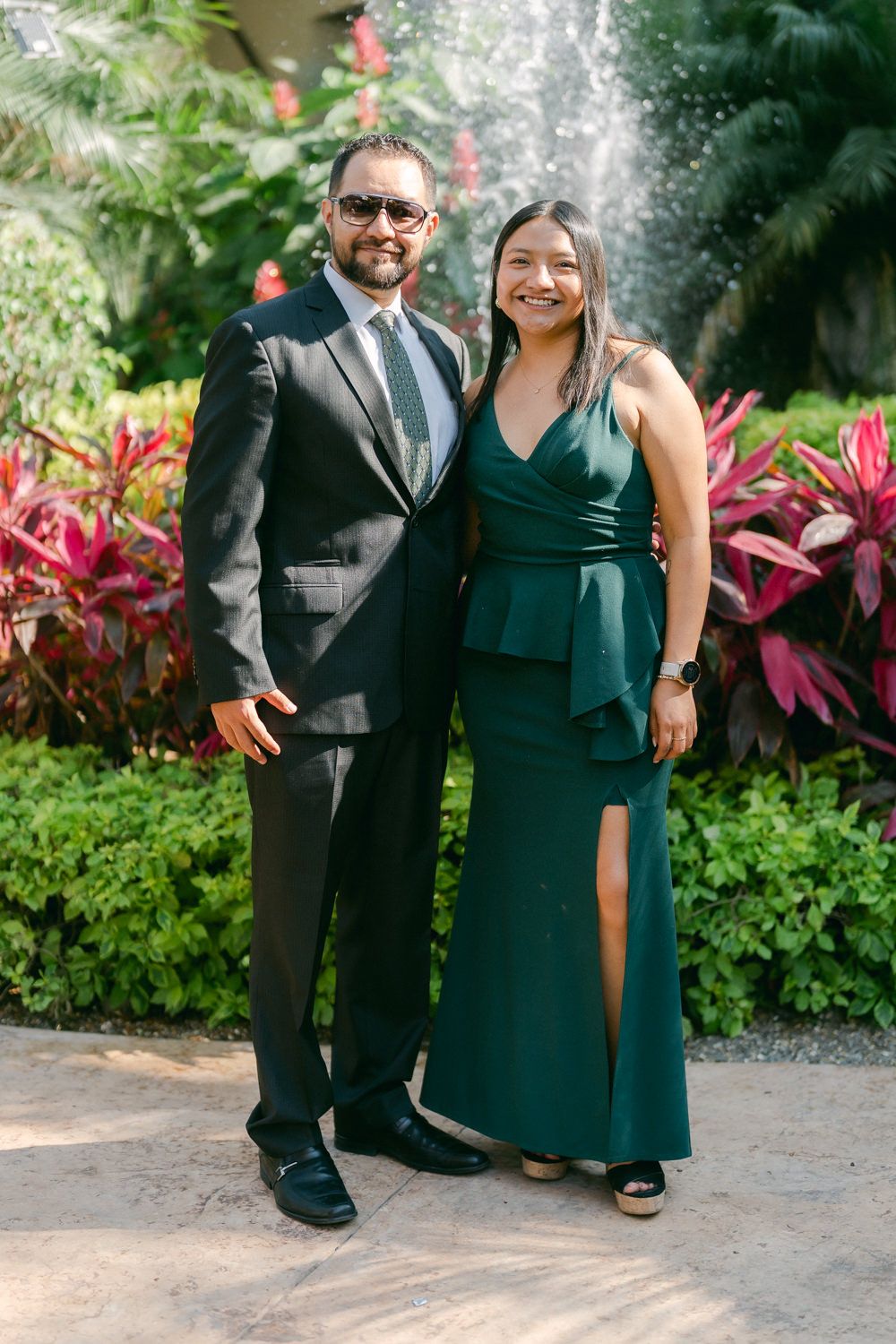 A couple in formal attire poses together in a garden setting with tropical plants and flowers in the background.