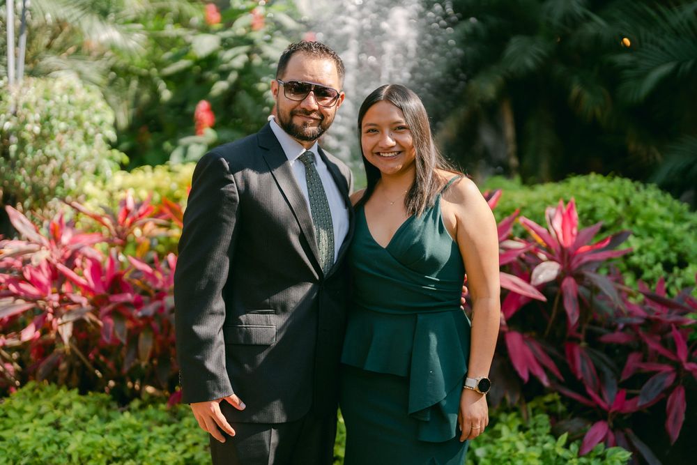 A couple in formal attire poses in a garden with vibrant pink foliage and tropical plants in the background.