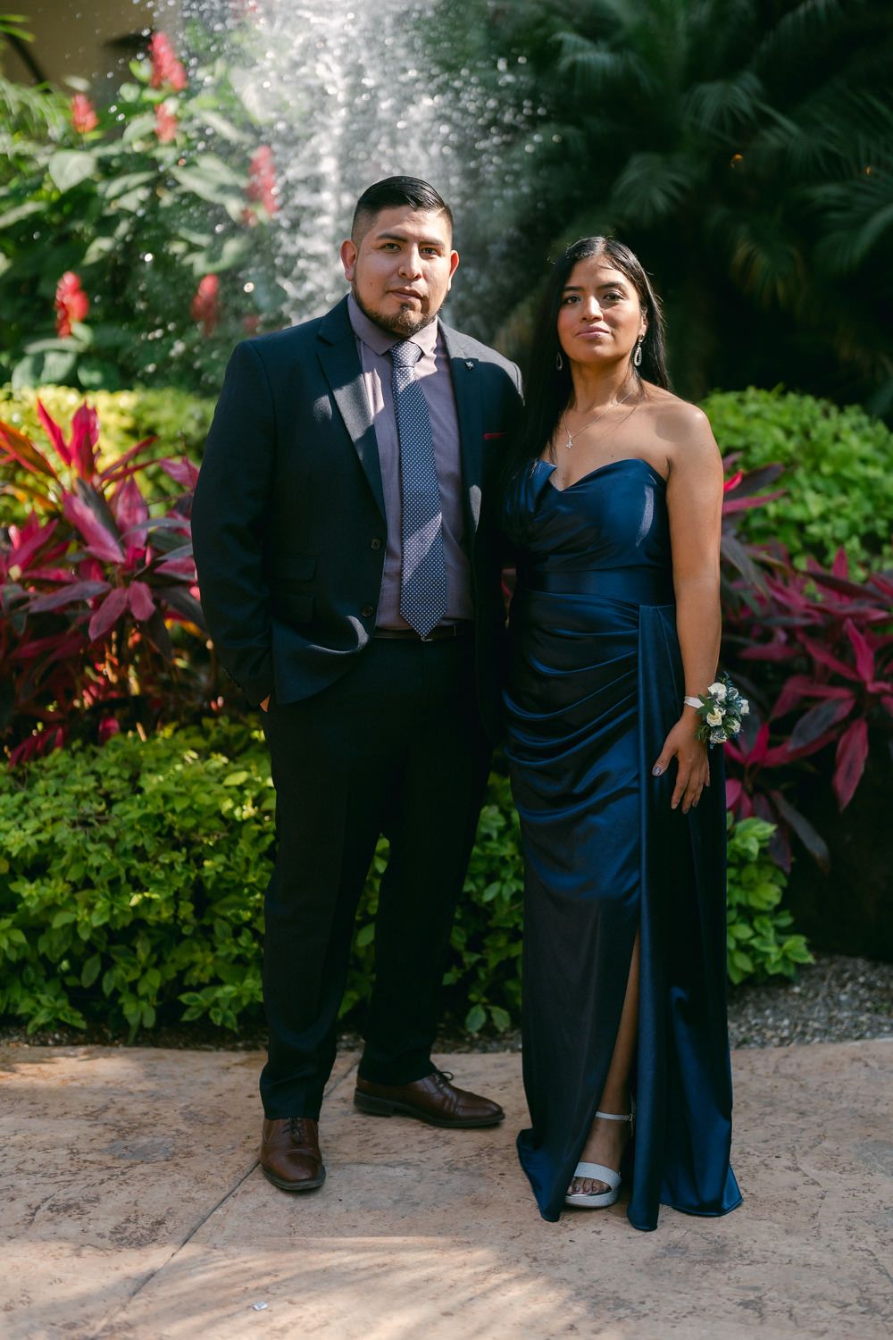 A couple in formal attire poses together in a garden setting with vibrant red foliage in the background.