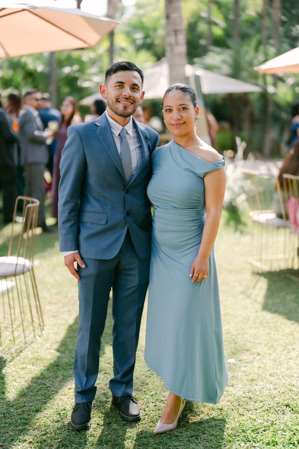 A couple poses together outdoors at an event, with the person in a navy suit and other in a light blue dress.