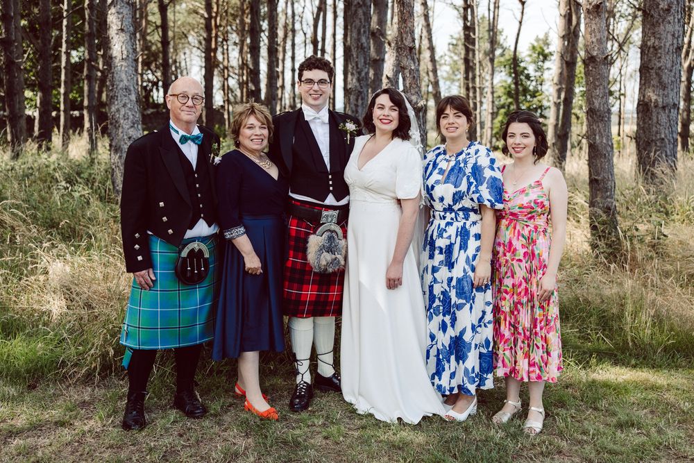 Wedding party members pose together in a wooded outdoor setting wearing formal attire and colorful dresses.