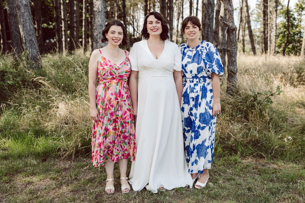Three women in floral and white dresses pose together outdoors in a natural wooded setting.