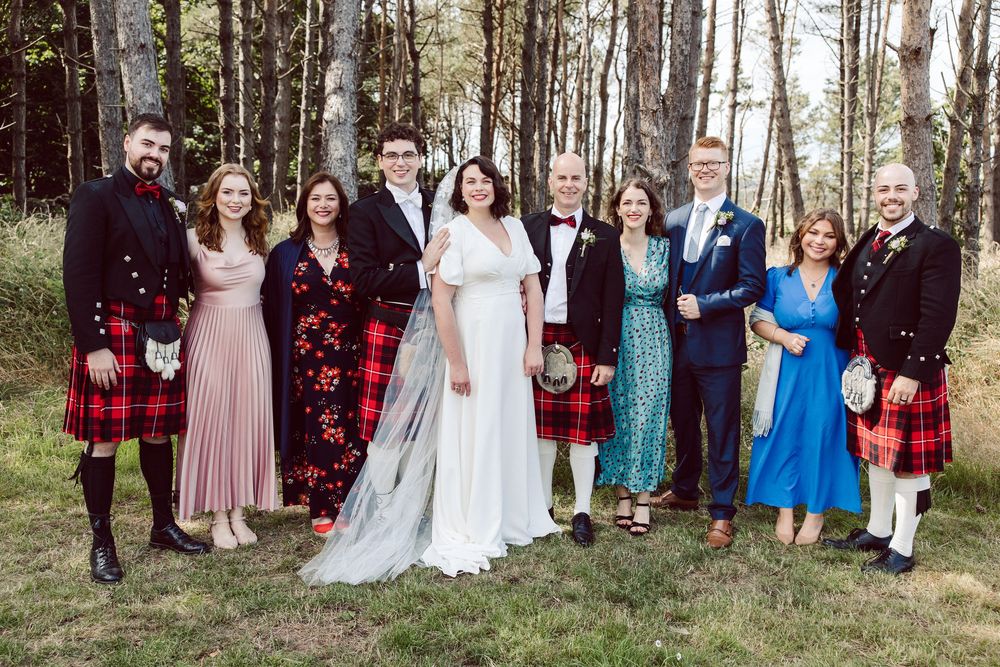 Wedding party poses outdoors in traditional Scottish kilts and formal attire among tall pine trees.