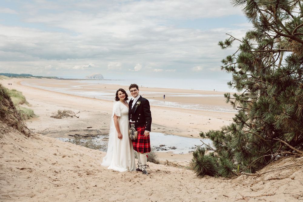 A couple stands on a sandy beach in traditional Scottish wedding attire with ocean and cloudy skies in the background.