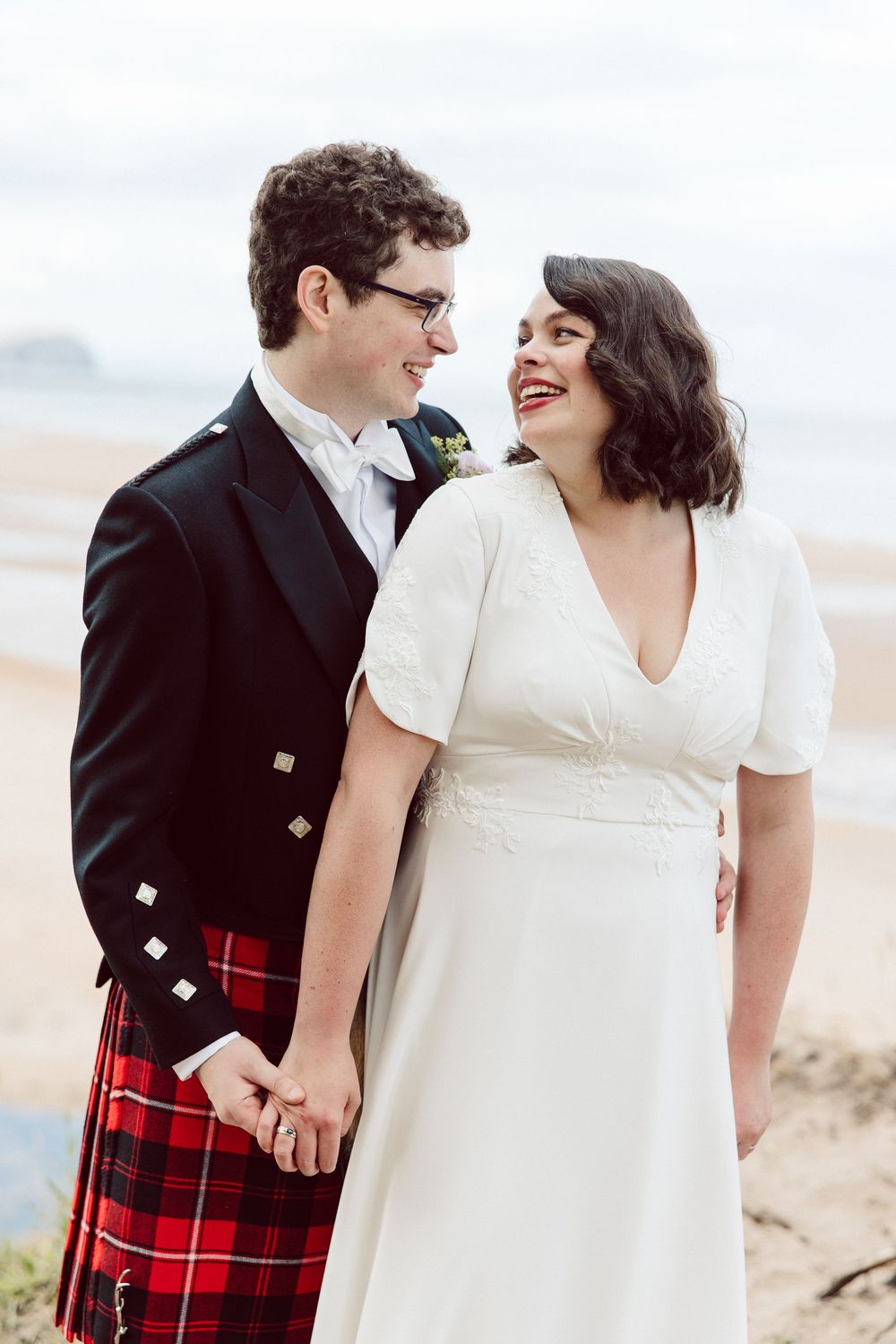 A couple shares a joyful moment on the beach, with the groom wearing a traditional Scottish kilt and black jacket.
