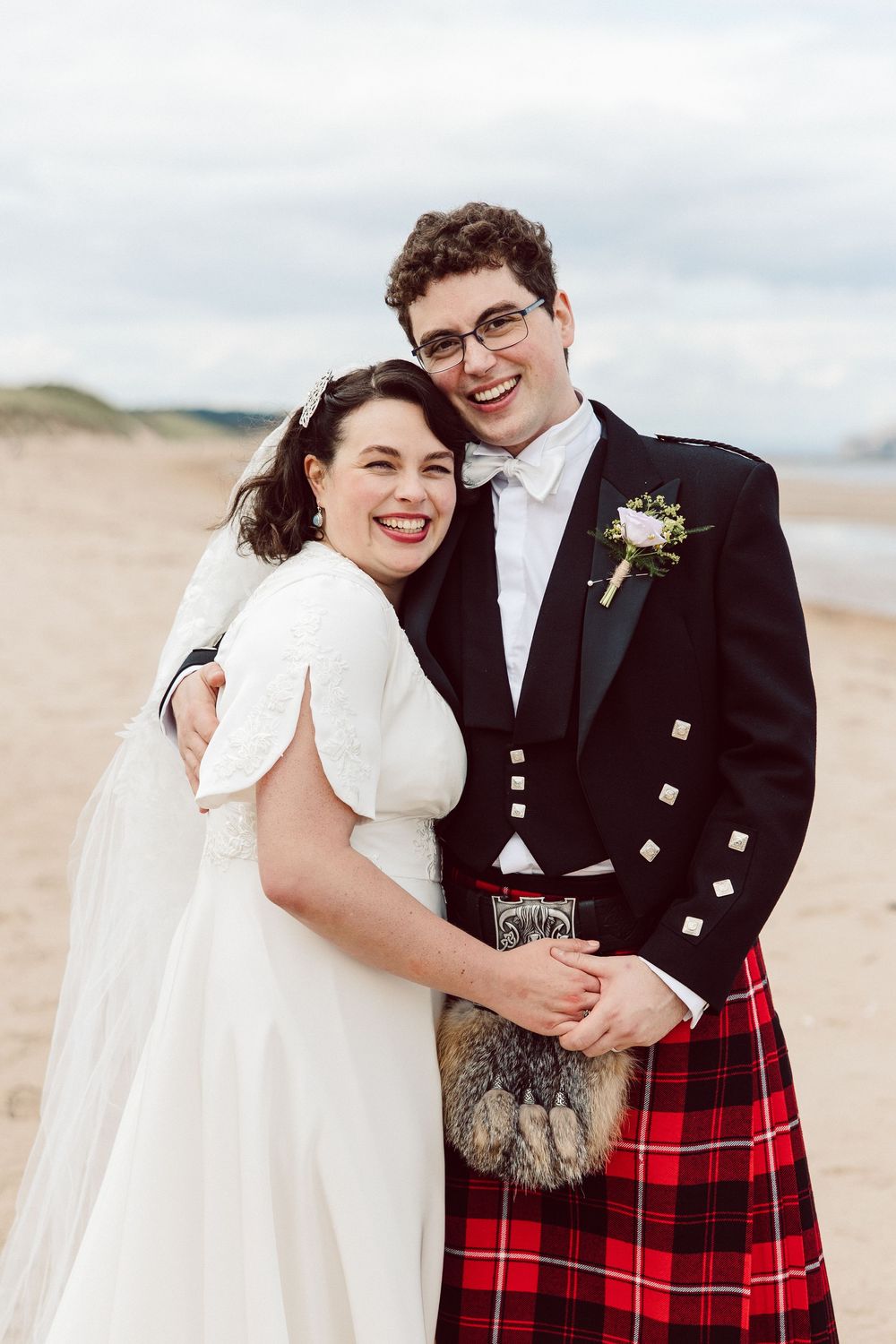 A bride and groom embrace on a sandy beach, with the groom dressed in a traditional red tartan kilt and formal jacket.