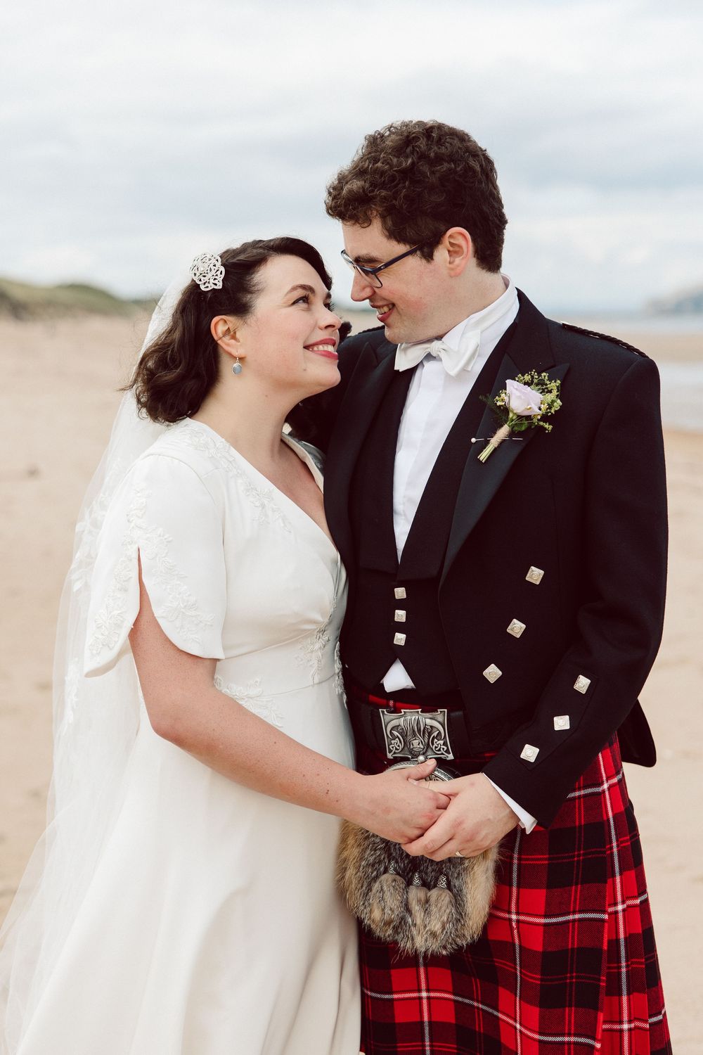 A couple in wedding attire share intimate moments on a beach, with the groom wearing a traditional Scottish kilt.