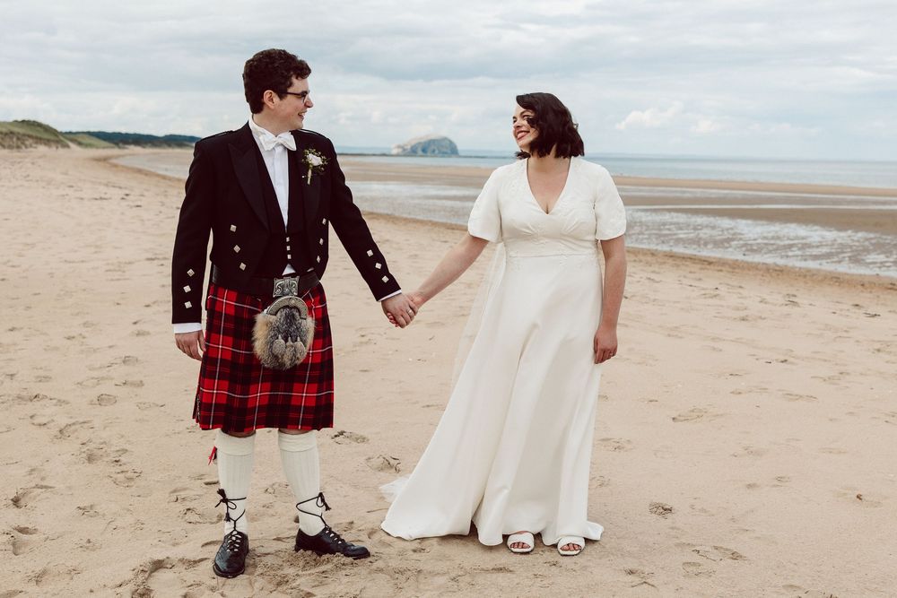 Couple in wedding attire holding hands while walking on a sandy beach with traditional Scottish kilt details.