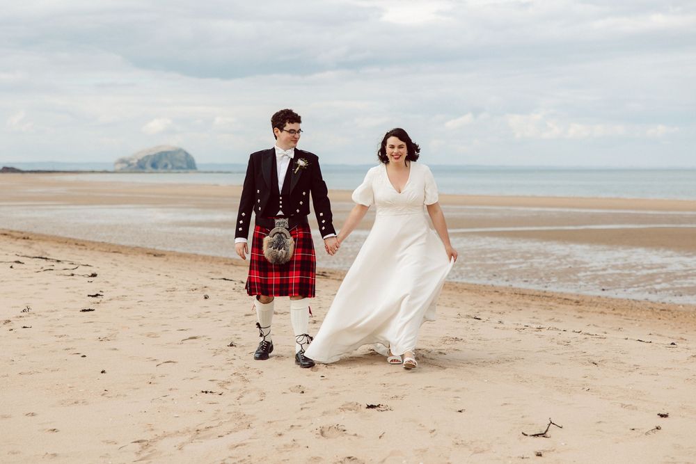 A couple in wedding attire walks along a scenic beach with mountains in the distance on their special day.