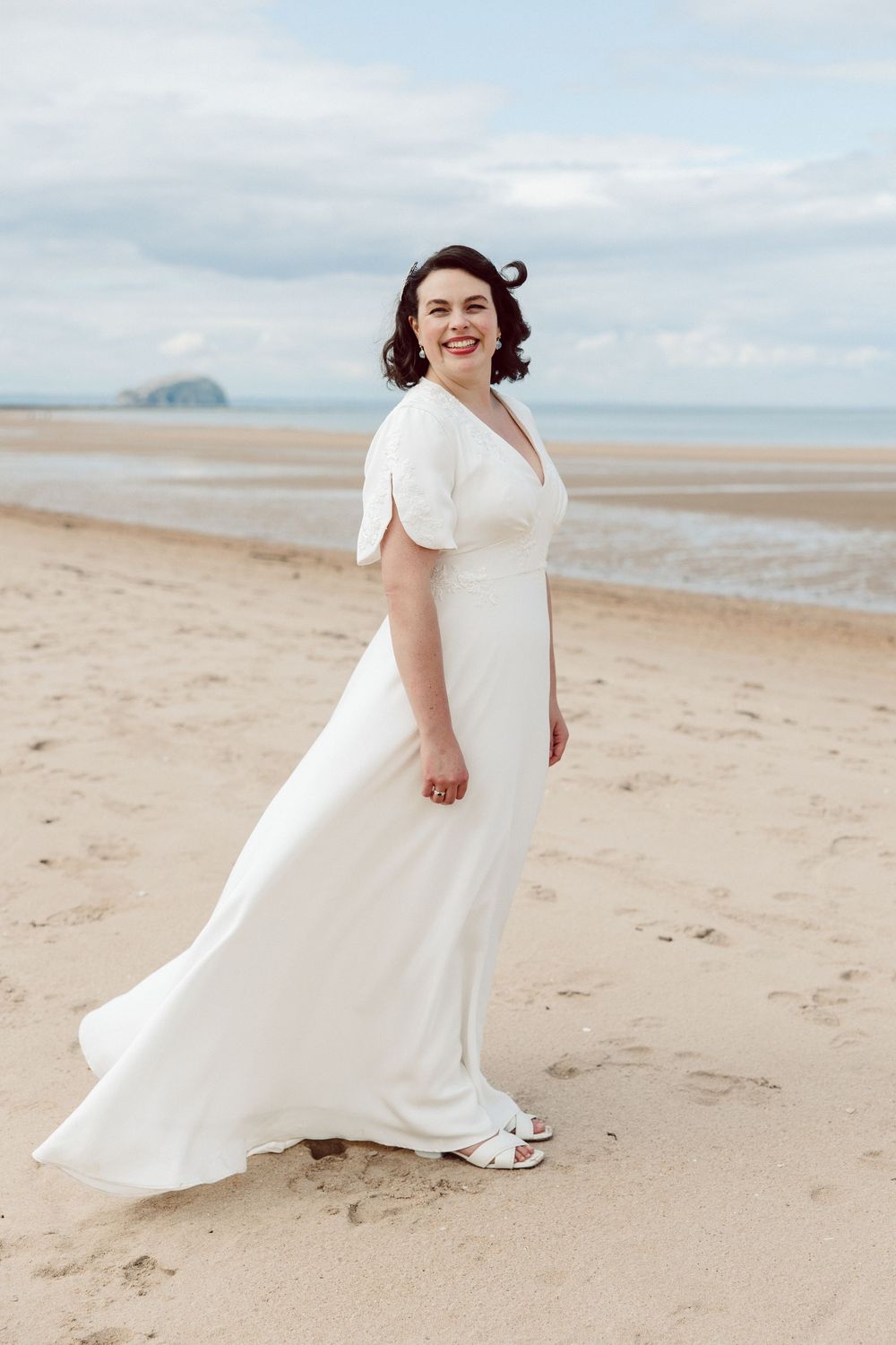 A person in a flowing white wedding dress twirls joyfully on a scenic beach with cloudy skies in the background.