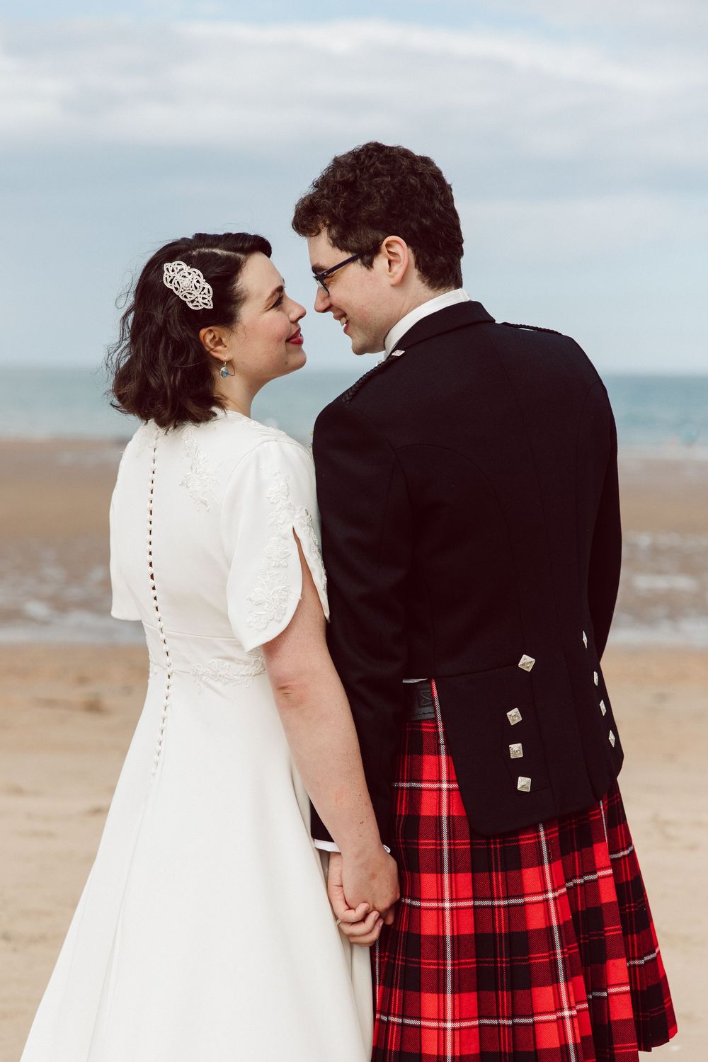Newlyweds share intimate moment on sandy beach with groom in Scottish formal wear and bride in white gown.