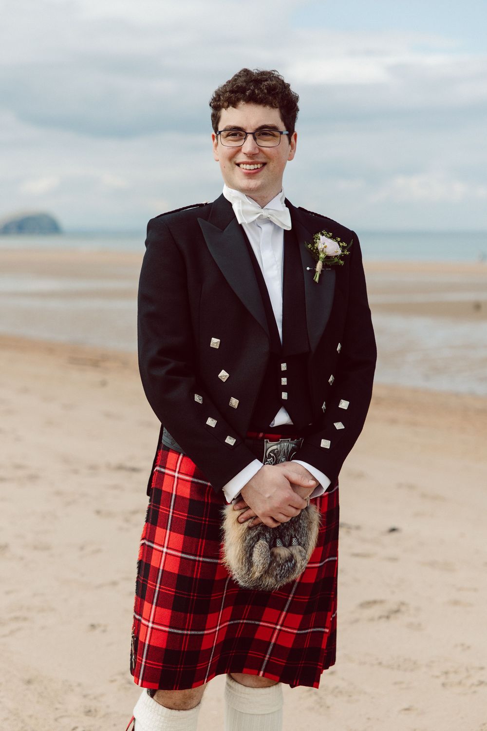Traditional Scottish wedding attire with red tartan kilt and formal black jacket photographed on a beach.