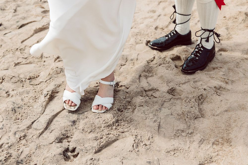 Close-up of bride and groom's feet on beach sand showing dress shoes and white sandals.