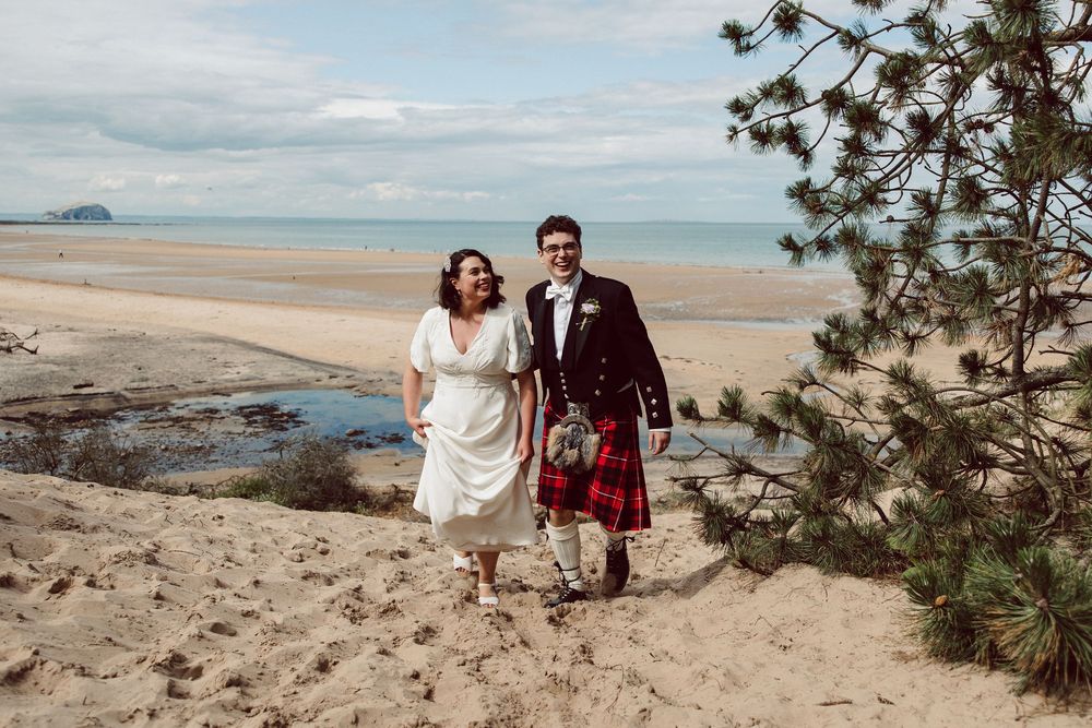 Couple walking along a scenic beach with pine trees and turquoise waters on a cloudy day.