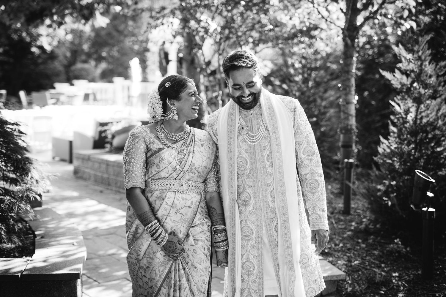 A couple in traditional Indian wedding attire walking together through a garden path in black and white photography.
