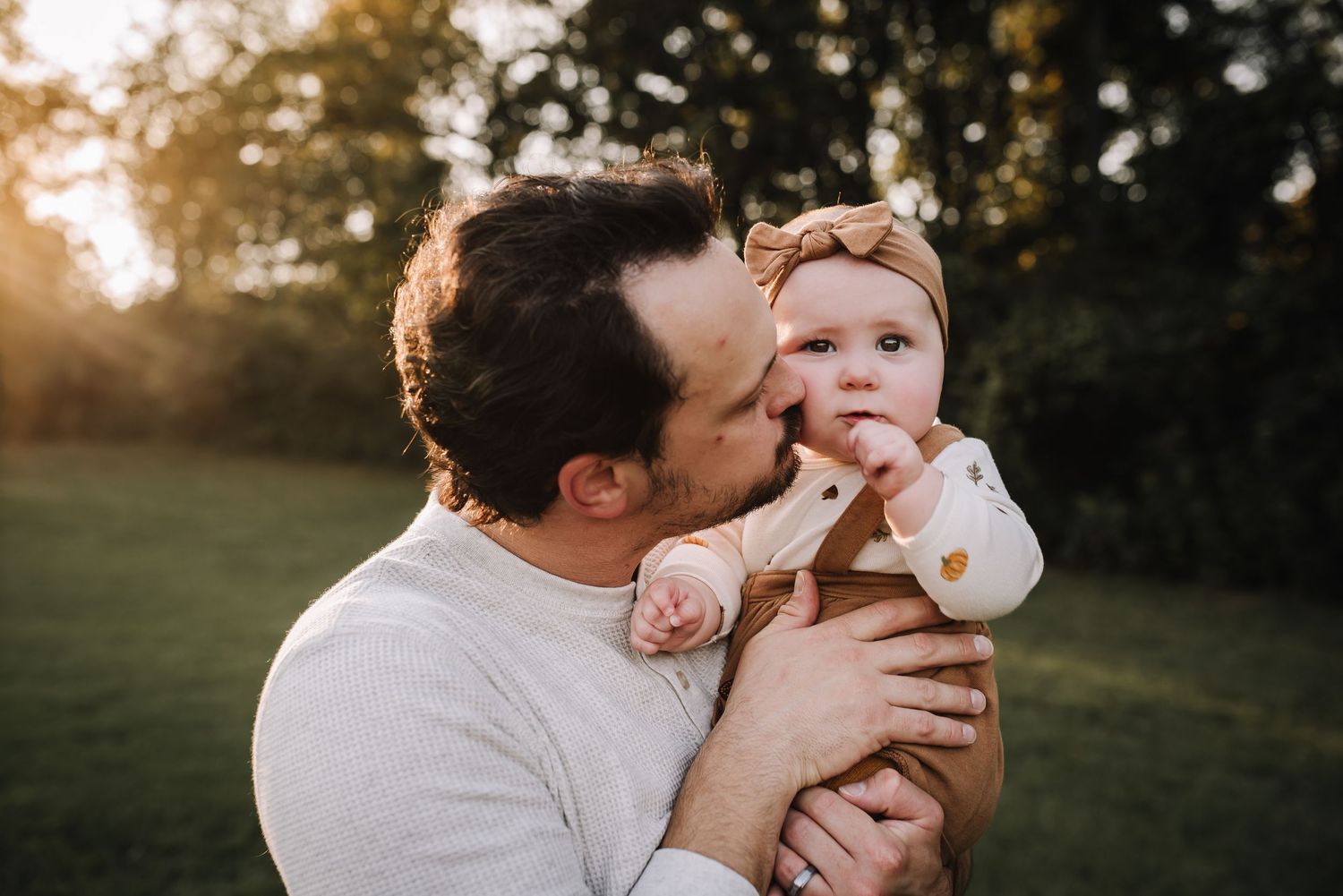 A parent kisses their baby during golden hour sunset in a grassy outdoor setting. Family photographer in Middle Tennessee capturing natural