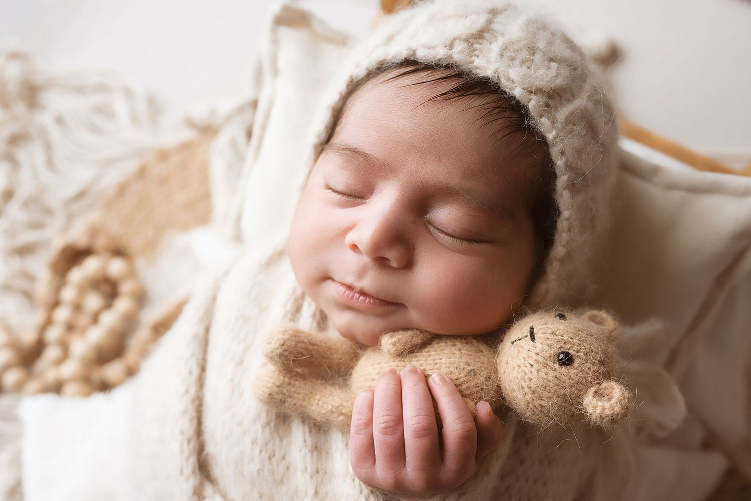 A sleeping newborn baby lies peacefully on cream-colored blankets with a small teddy bear nearby.