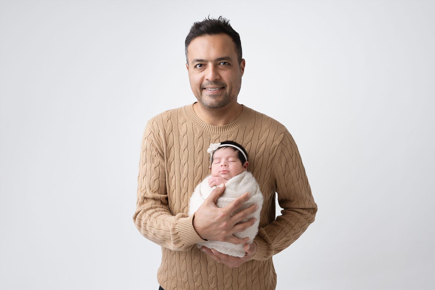 A parent holds a small baby while wearing a beige sweater against a white studio backdrop.