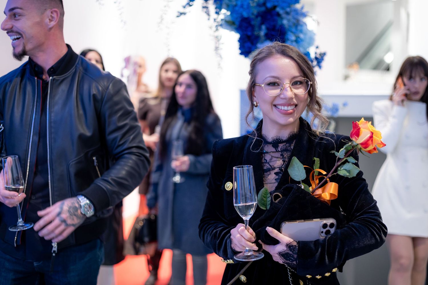 Woman smiling at event holding champagne glass and roses in black velvet outfit.
