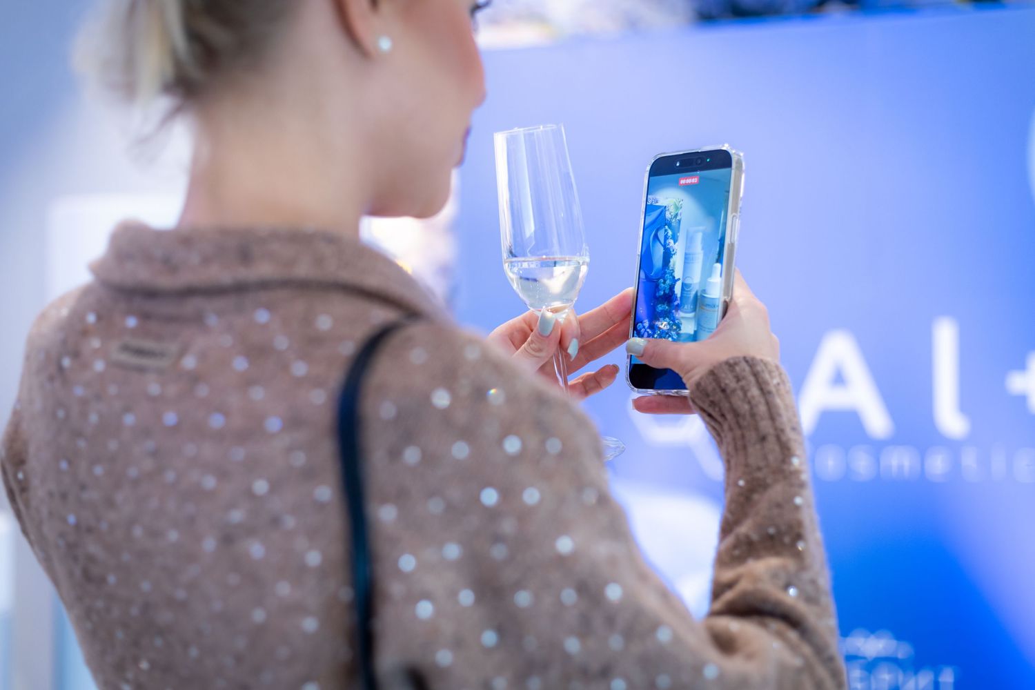 Woman holding champagne glass and smartphone photographing display at cosmetics event.