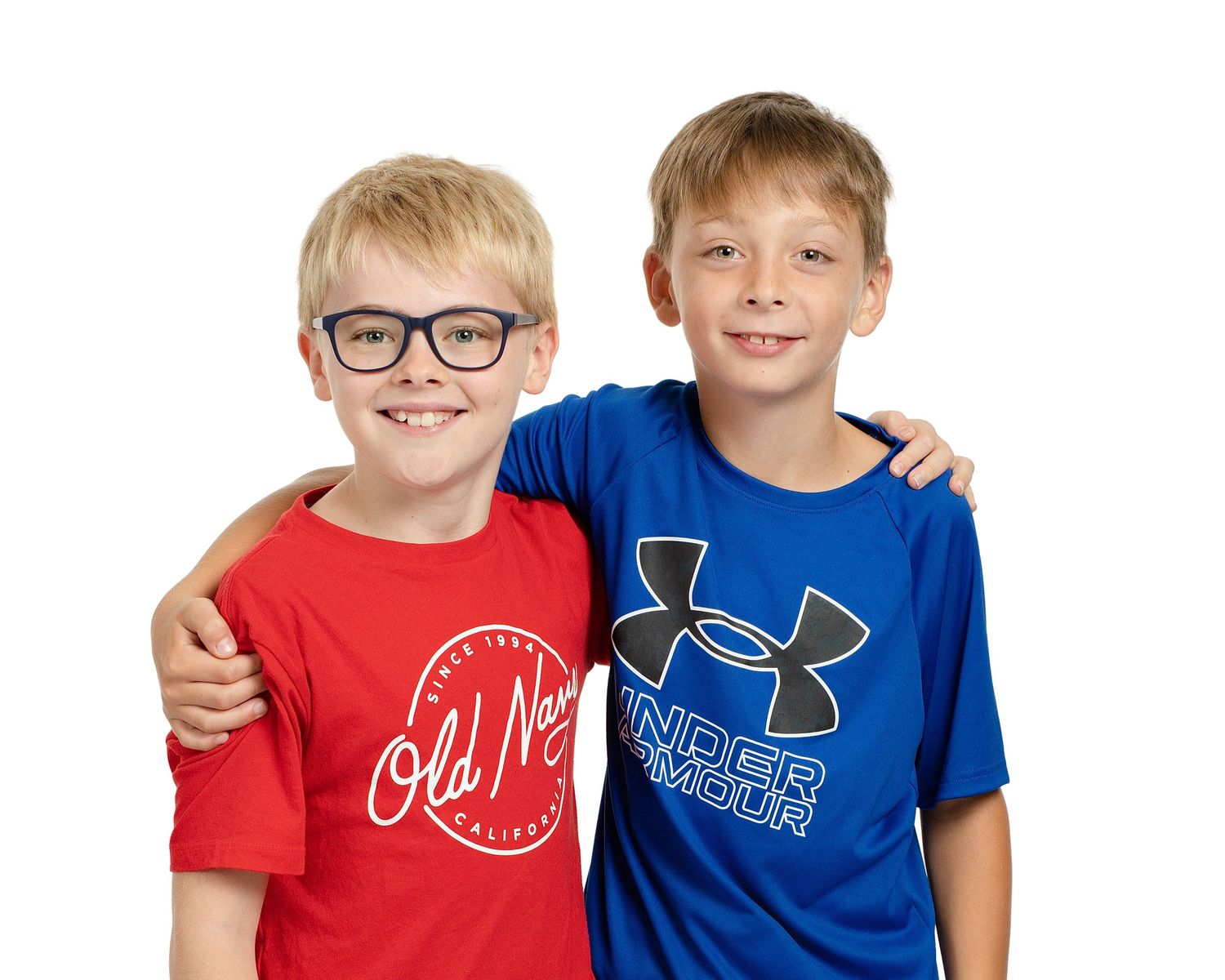 Two young boys wearing glasses and sports brand t-shirts smile together with arms around each other's shoulders.