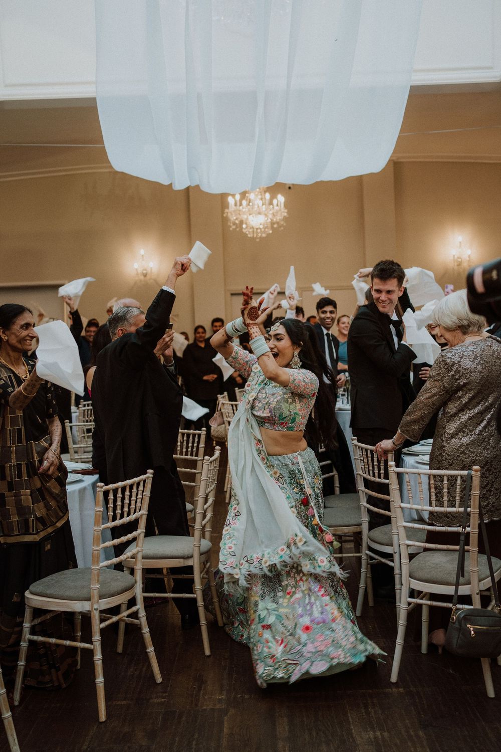 Wedding guests toss white napkins in the air during a celebratory reception moment in an elegant ballroom setting.