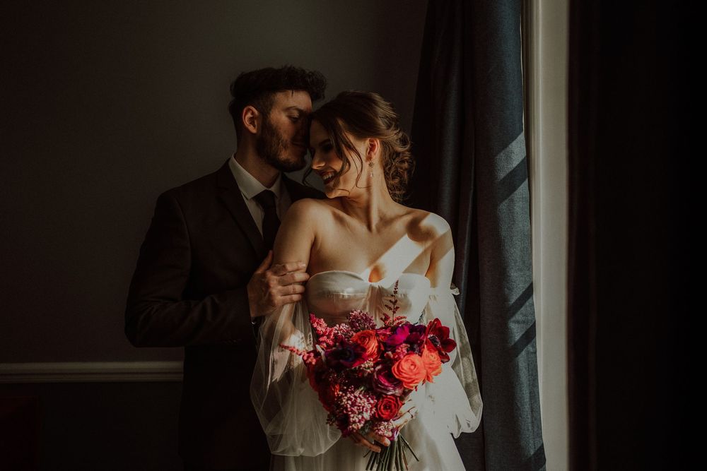 A couple in wedding attire embraces near a window in moody, dramatic lighting at bourne and hollingsworth