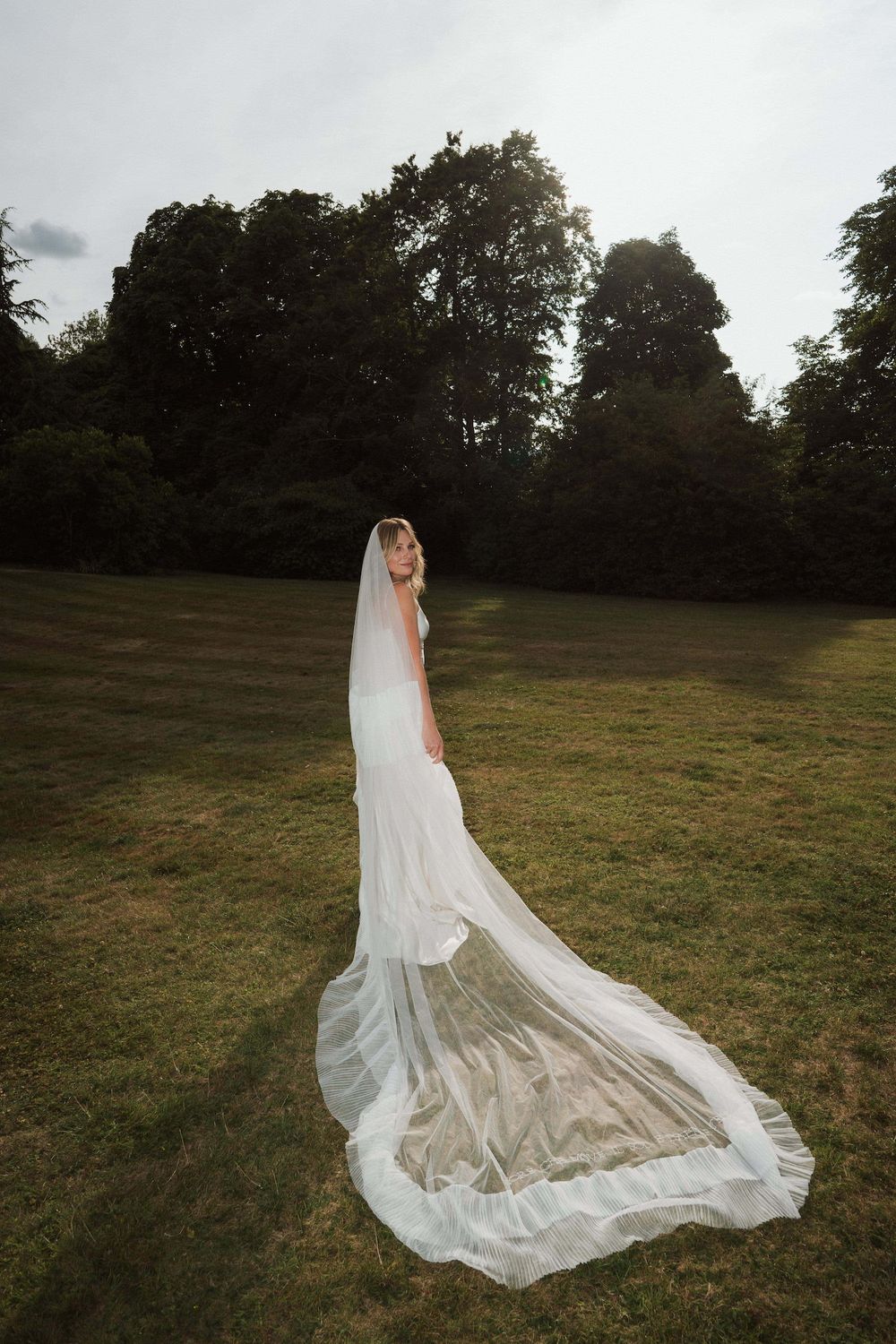A flowing white wedding dress with a long train spreads elegantly across a grassy hillside at dusk at Sudeley Castle, Cotswolds