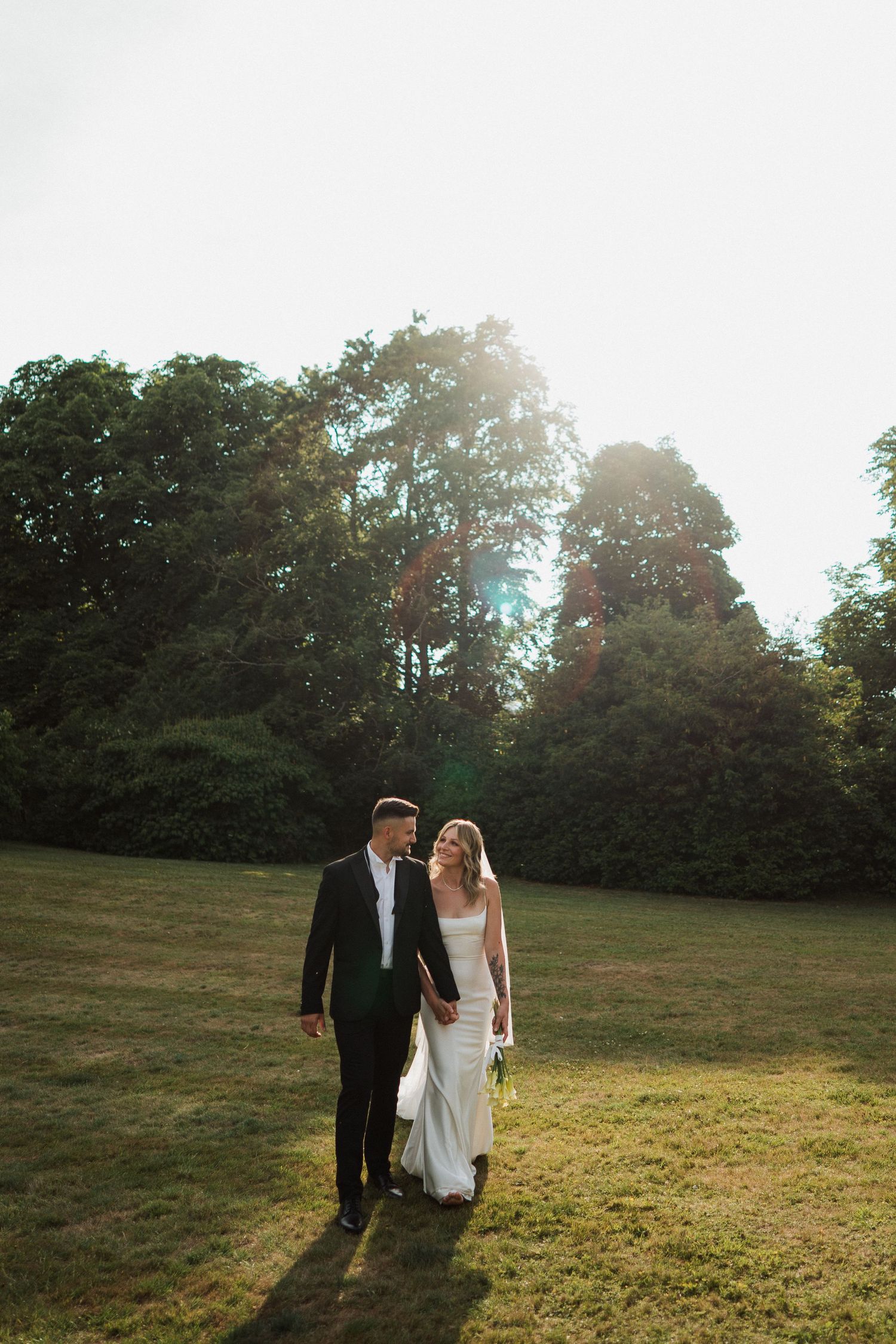 A couple walks together on a sunlit grassy hill with trees silhouetted in the background during golden hour at Sudeley Castle, Cotswolds