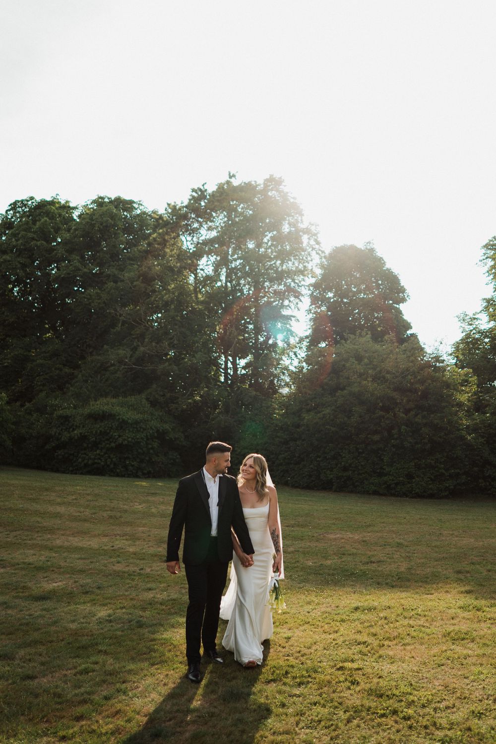 A couple walks together on a sunlit grassy hill with trees silhouetted in the background during golden hour at Sudeley Castle, Cotswolds