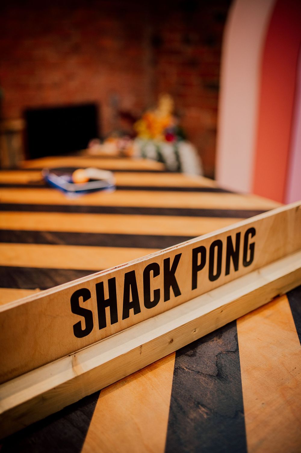 Close-up shot of a wooden Shack Pong game table with black lettering against a warm-lit brick wall background.