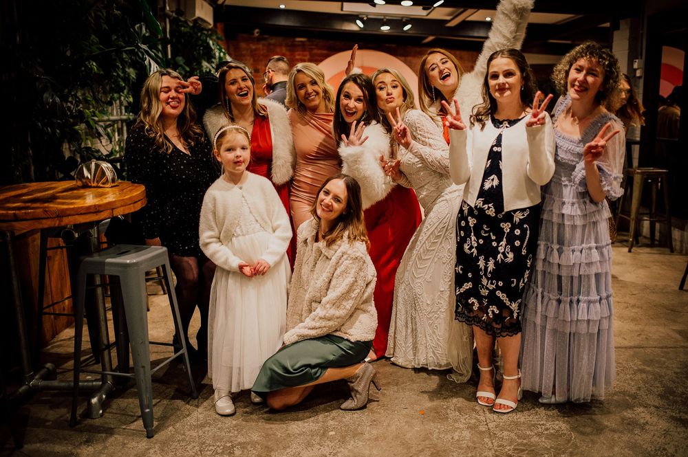 A group of people pose together at a party venue wearing an assortment of dresses and festive attire.